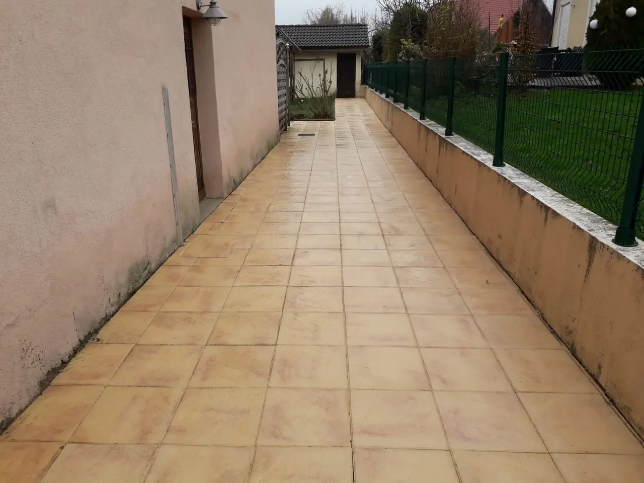 Allée en dalles ocre qui longe la façade d’une maison jusqu’à un abri de jardin, muret et grillage vert à droite.